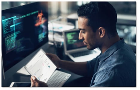 Man reading a tablet screen while sitting in front of a large desktop monitor