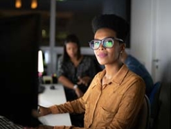 Woman in large glasses and a brown long sleeve shirt working at a monitor 