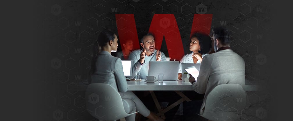 People in a conference room around a laptop with a large red W behind them