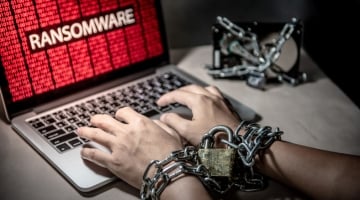 Hands chained with a padlock typing on a laptop showing a ransomware attack warning; symbolizing data encryption and cyber extortion.